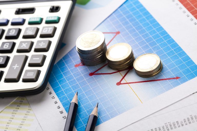 Em cima da mesa há uma calculadora, dois lápis, várias moedas empilhadas em cima de um gráfico