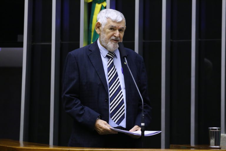 Deputado Luiz Couto fala ao microfone. Ele é branco, tem o cabelo branco, usa um terno preto com camisa clara e gravata listrada e segura um papel