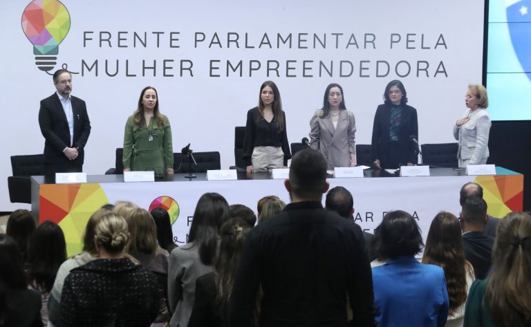 Várias pessoas estão em pé atrás de uma mesa, e outras pessoas estão na plateia também em pé